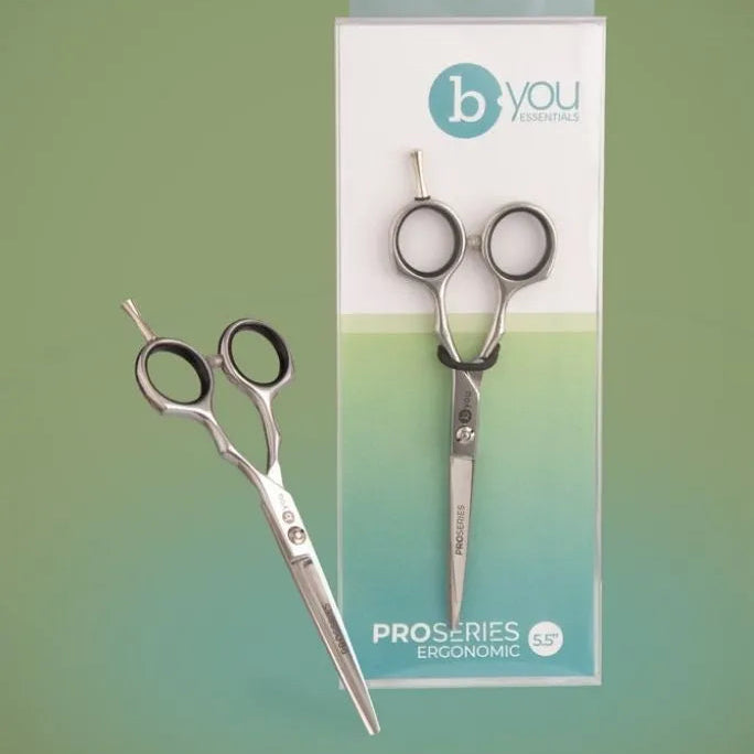 A pair of silver ProSeries Ergonomic scissors by bYOU Haircare, shown both loose and inside its retail packaging against a solid sage green background.