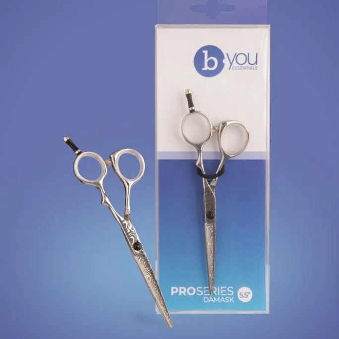 A pair of silver professional hair scissors shown next to their retail packaging on a blue gradient background. The brand 'b you' is visible on the box.