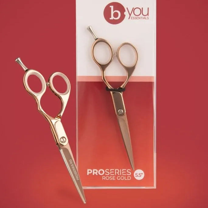 Rose gold hair shears scissors shown next to their retail packaging on a solid red background. The brand 'b you' is visible on the packaging.
