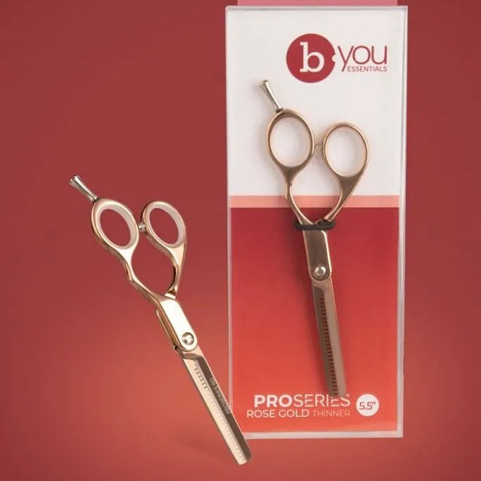 A pair of gold-toned professional hair shears shown next to their retail packaging on a solid dark red background. The packaging is white and red with the brand logo 'b you' and the text 'PROSERIES ROSE GOLD TINNY'.